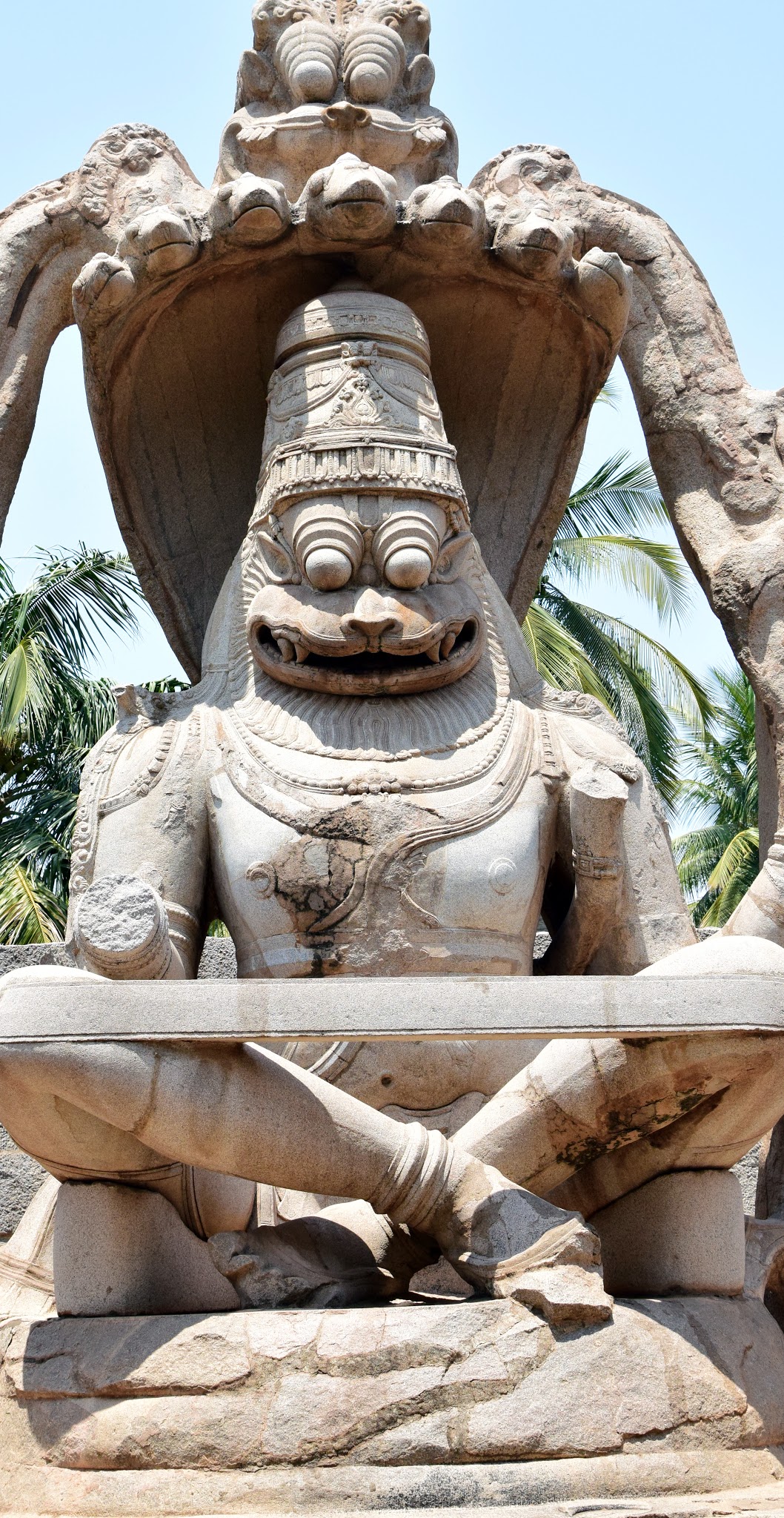 Photograph of the massive monolithic Lakshmi Narasimha stone statue at Hampi — a fierce lion-faced deity seated cross-legged under a hooded canopy, palm trees behind